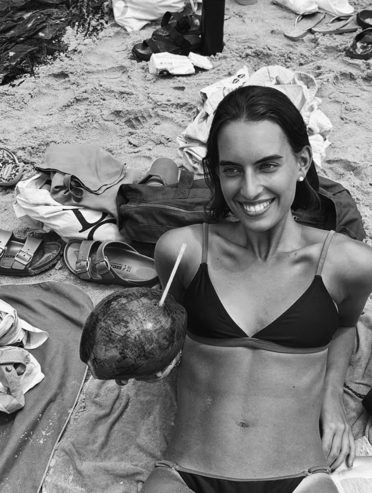 Smiling woman in bikini holding coconut at beach, surrounded by shoes and personal items on sand, black and white photo